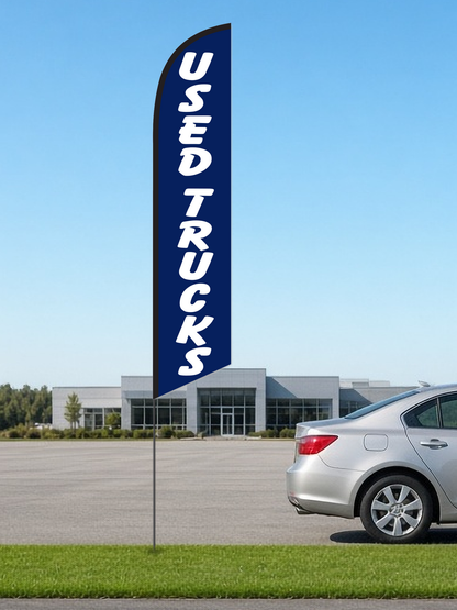 Used Trucks swooper feather flag, white USED TRUCKS lettering on navy blue, black pole sleeve, on a pole in the grass strip of a dealership lot beside a silver sedan