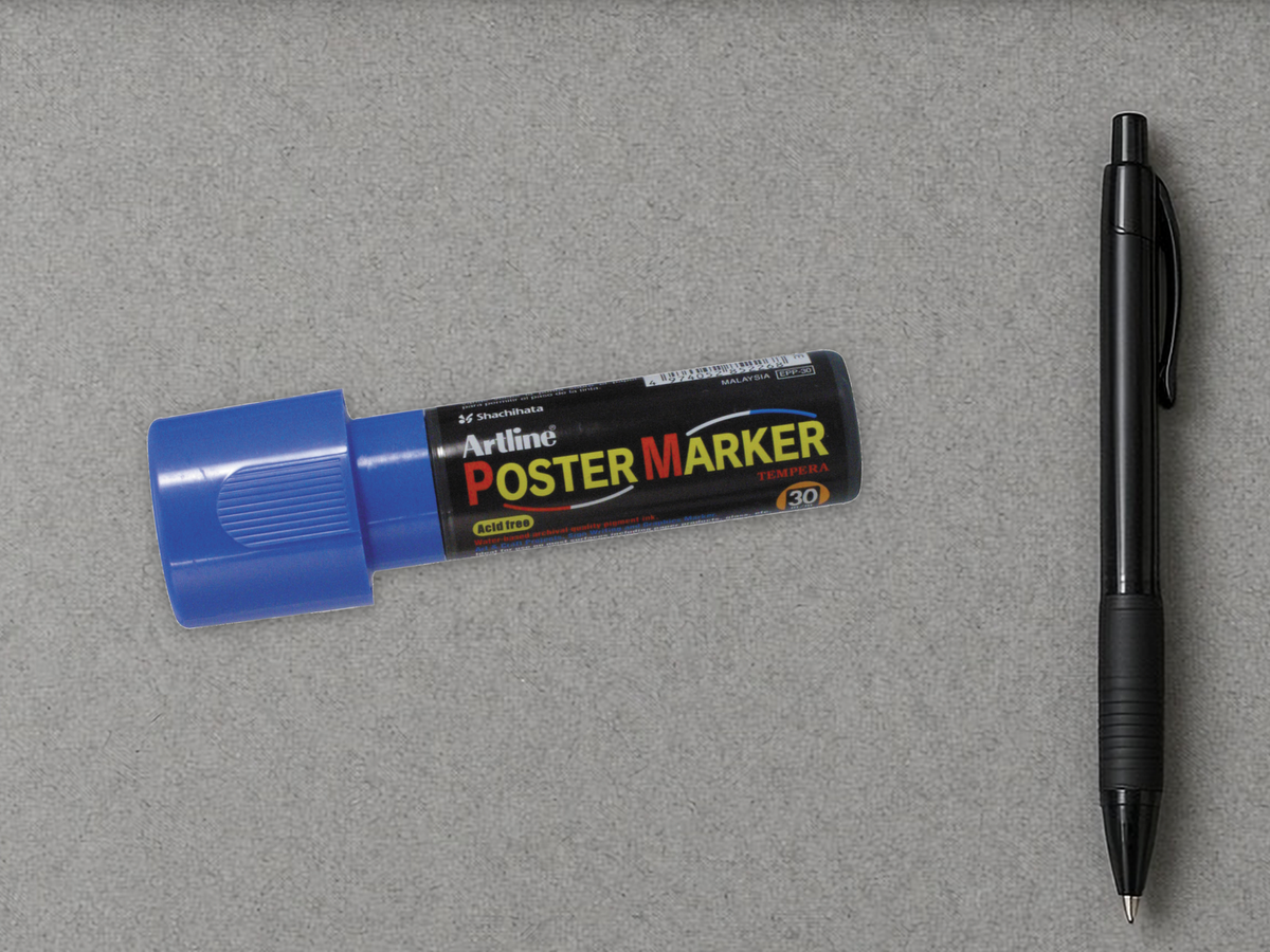 Blue Artline Poster Marker 30 mm wide-tip paint marker lying on a gray parts counter beside a pen